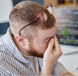 Man with eye strain rubbing eyes at desk, a common trigger for eye strain migraines