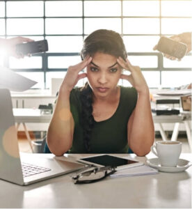 Overwhelmed woman at desk clutching her head—illustrating the impact of sensory overload headaches in a modern workspace.
