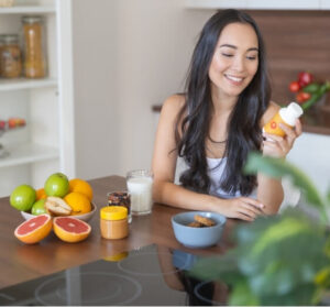 A smiling woman holding a supplement bottle while sitting at a table with fruit and juice – migraine supplements for natural support.